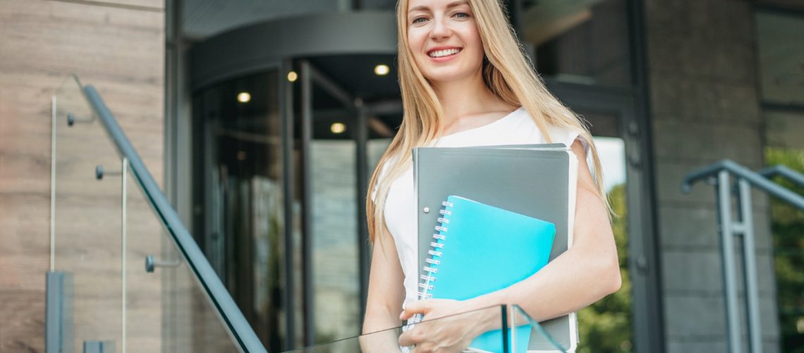caucasian-student-girl-holds-folders-notebooks-hands-smiles-background-university-building-copy-space